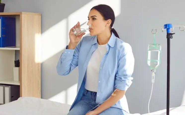 Woman drinking water beside an IV stand at home